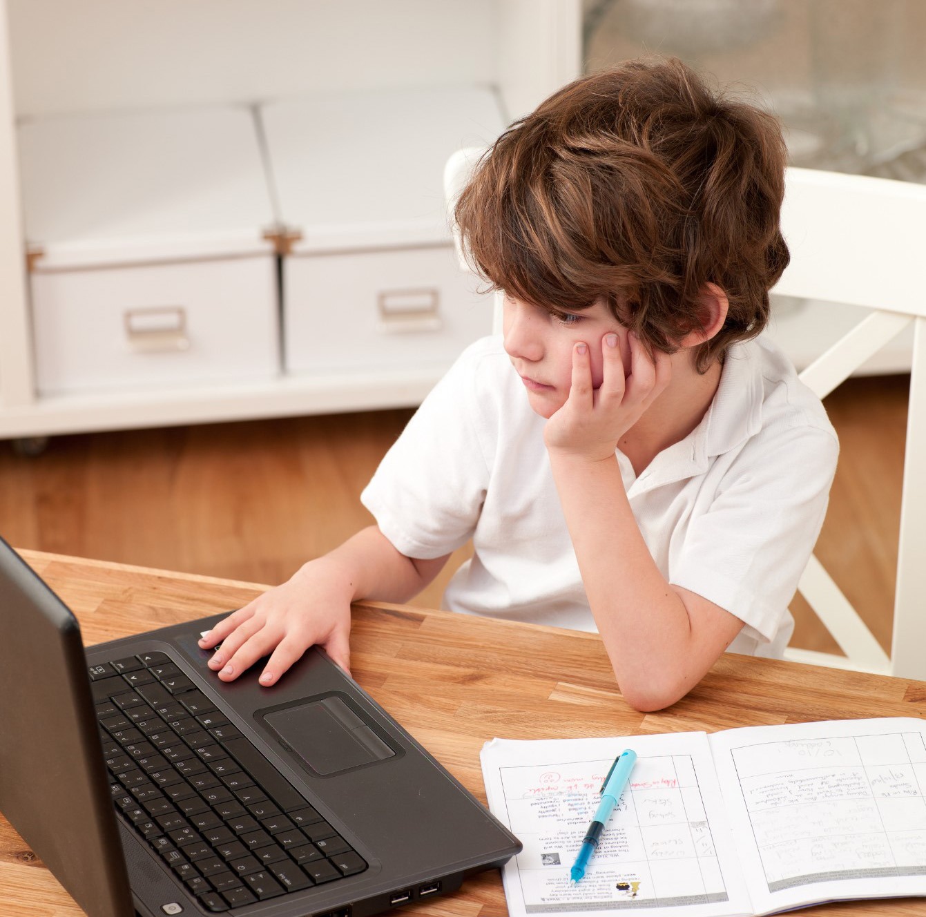 A focused child rests his chin on his hand while using a laptop, with schoolwork and a pen beside him on the table.