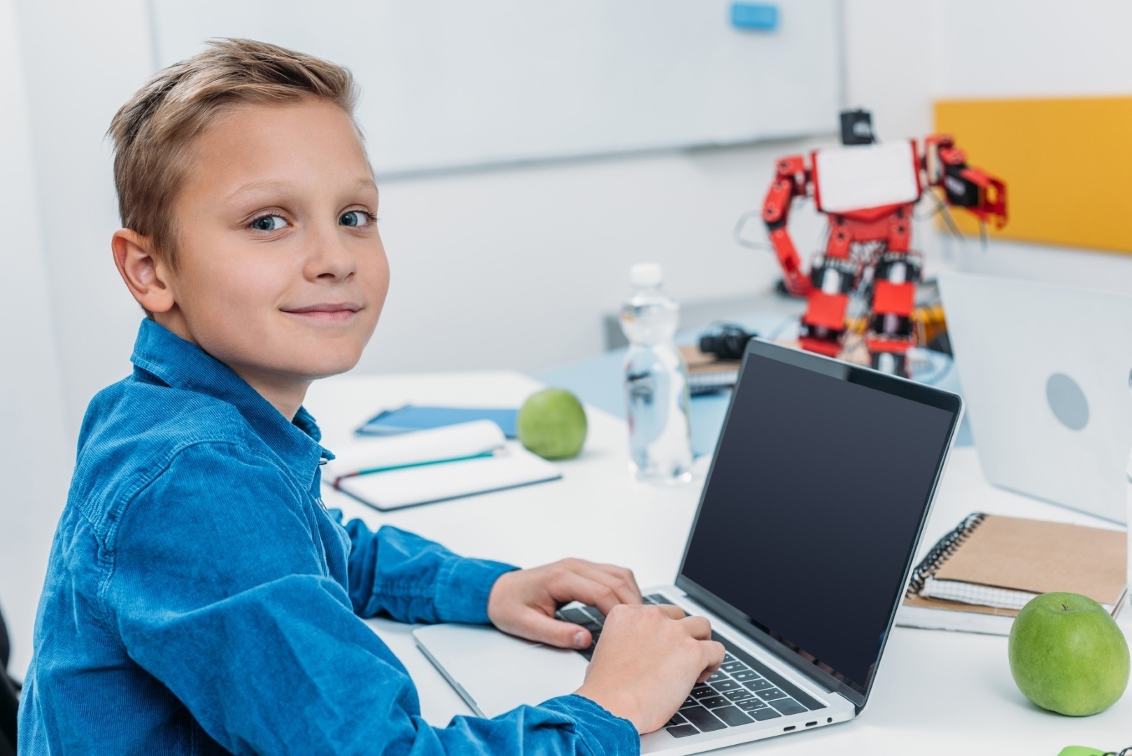 A young boy in a blue shirt sits at a desk, working on a laptop. A robot toy and green apples are visible, indicating a creative learning environment.