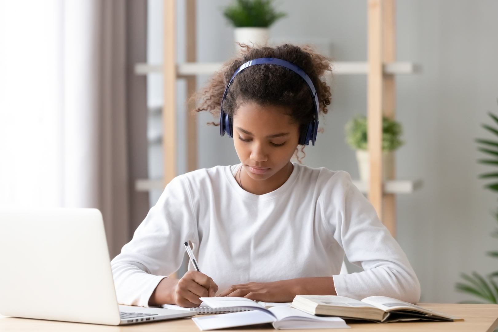 Student wearing headphones studies at a desk, focused on writing in a notebook while a laptop and open book are nearby.