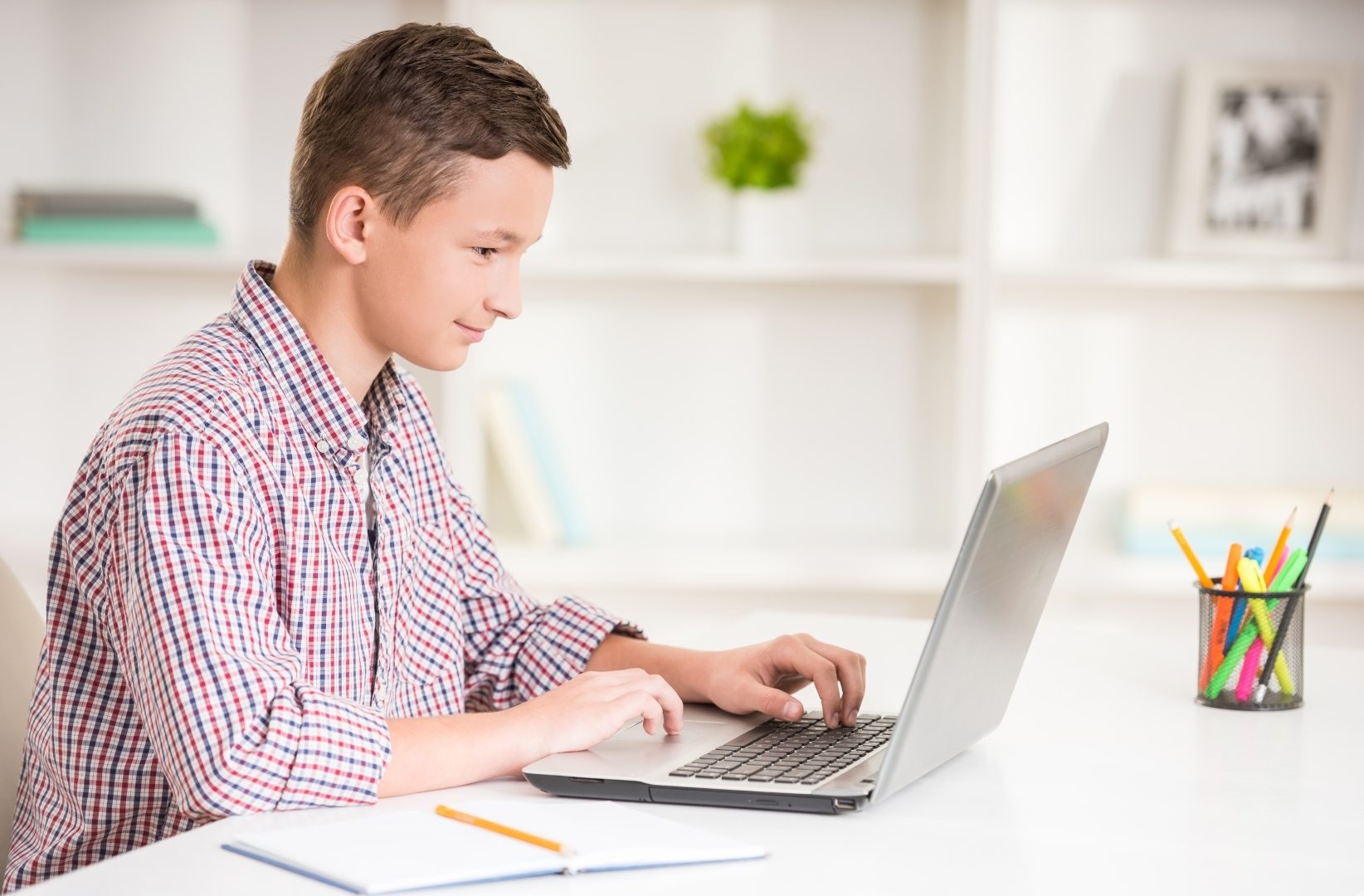 Boy in a plaid shirt focuses on a laptop in a bright, organized workspace. A cup of colorful pens and a notebook are nearby, suggesting study or homework.