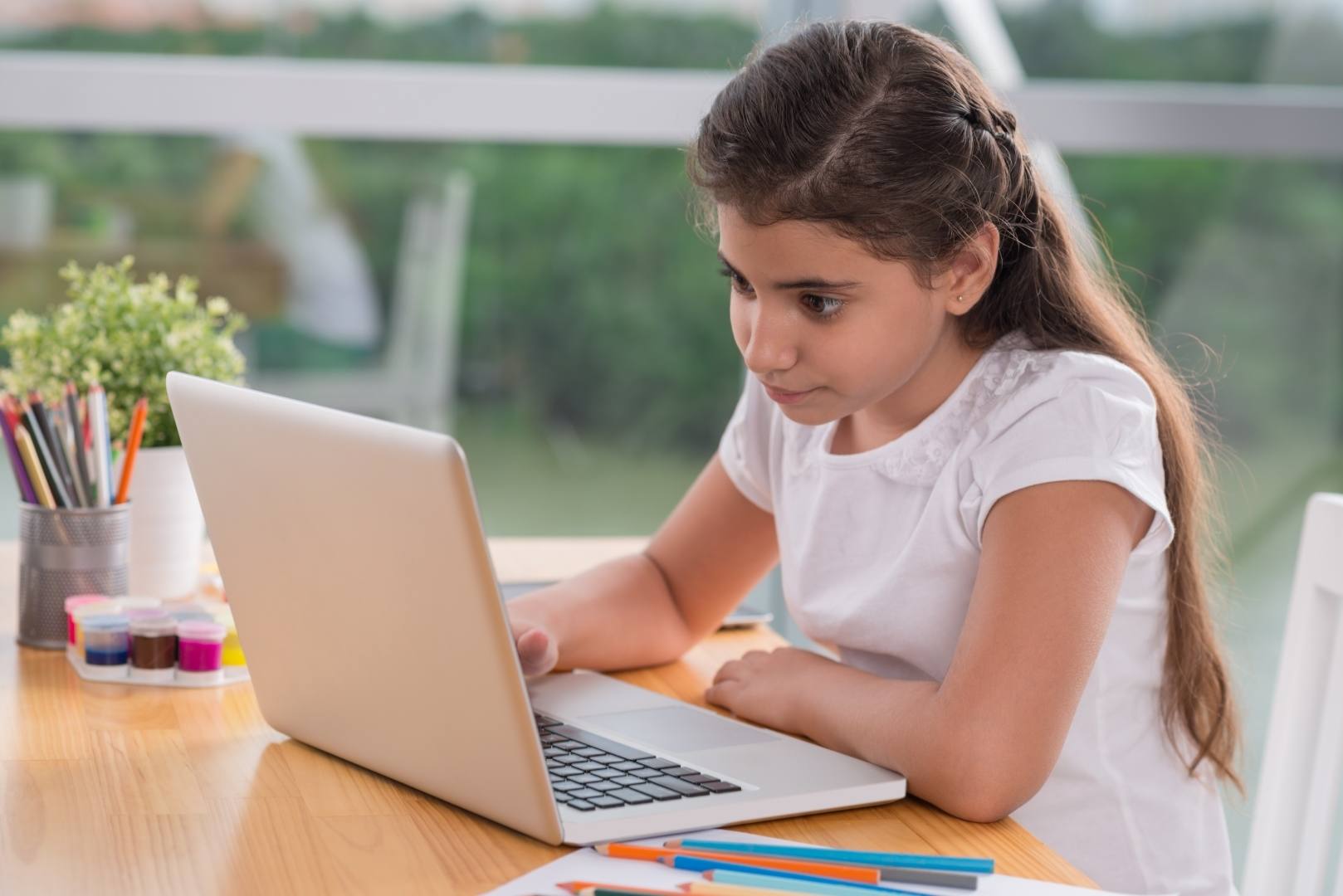 A young girl in a white shirt focuses intently on a laptop, surrounded by colorful art supplies and plants, suggesting a creative learning environment.