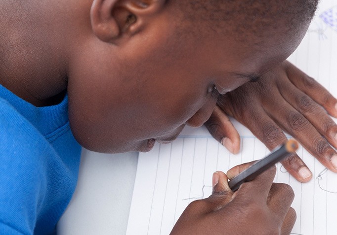 Child concentrating on drawing while lying on a desk, with a pencil in hand and lined paper beneath, highlighting focus and creativity.