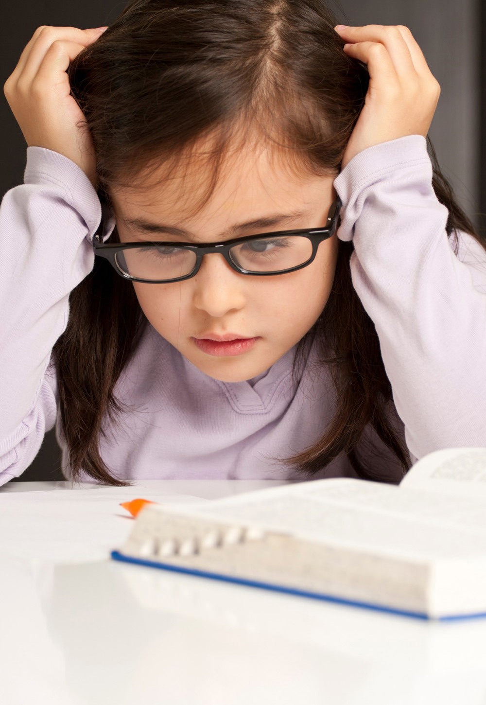 Young girl with glasses looks stressed while studying a book, resting her head in her hands. This image conveys the challenges of learning.