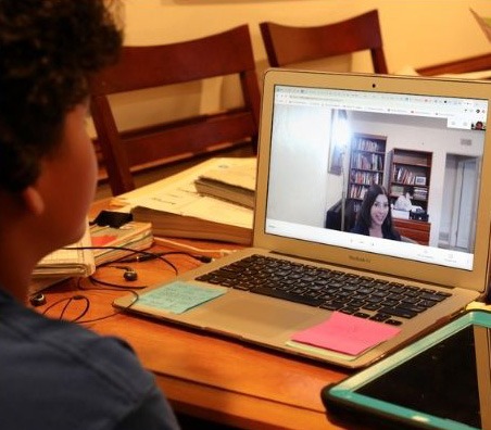 A child participates in a video call with a teacher, focused on a laptop surrounded by study materials, highlighting remote learning.
