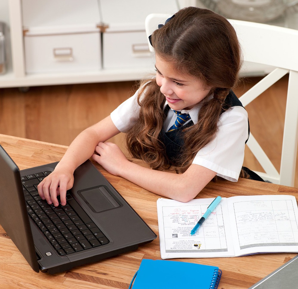 A school-aged girl, wearing a uniform, happily engages with a laptop at a wooden desk, surrounded by notebooks and a pen, focusing on her studies.