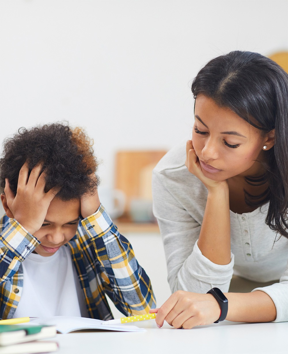A child struggles with homework, showing frustration, while an attentive adult offers support and guidance in a bright, calm environment.