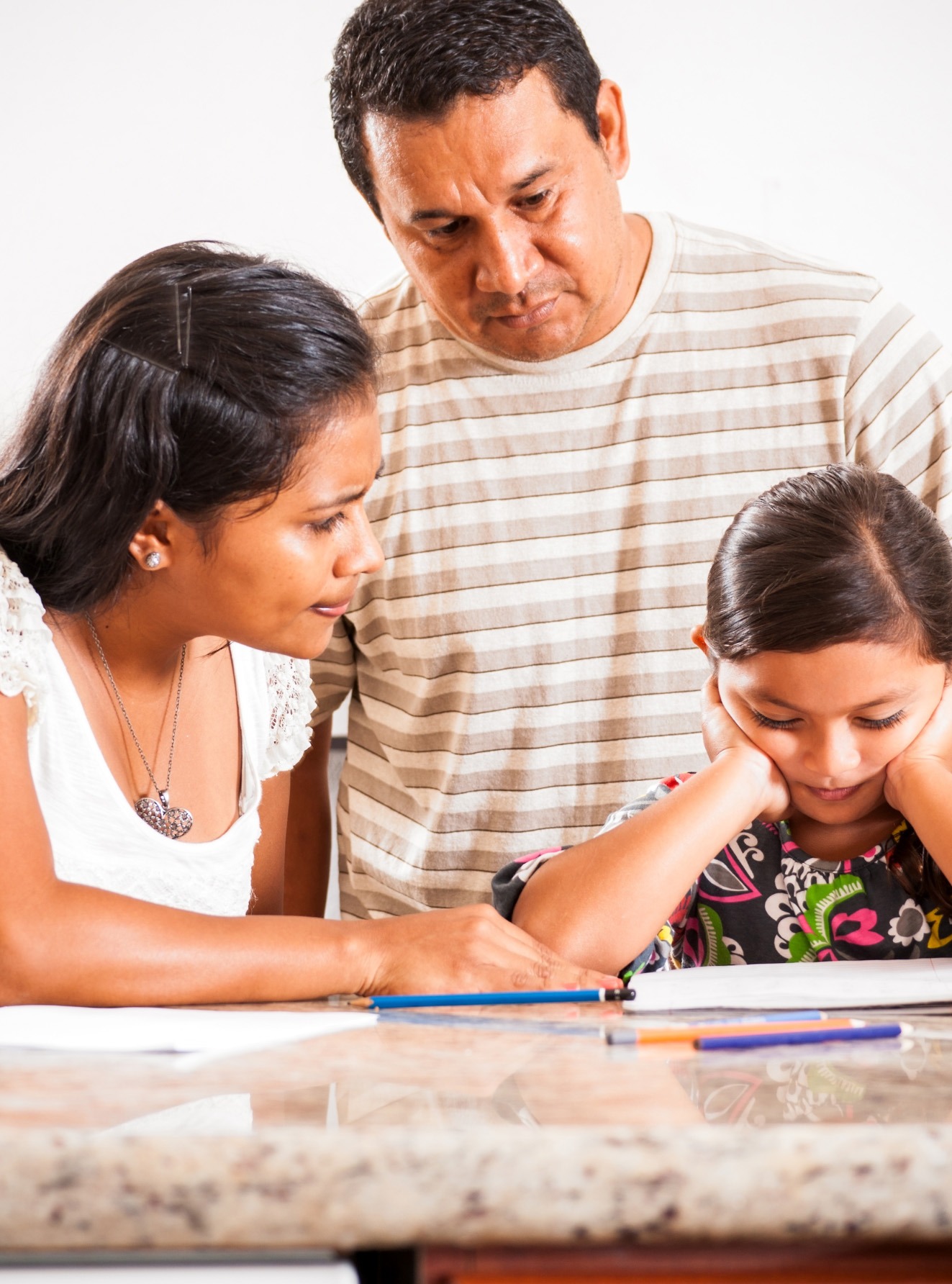 A concerned mother and father help their daughter with homework, as she rests her chin on her hands, showing signs of frustration.