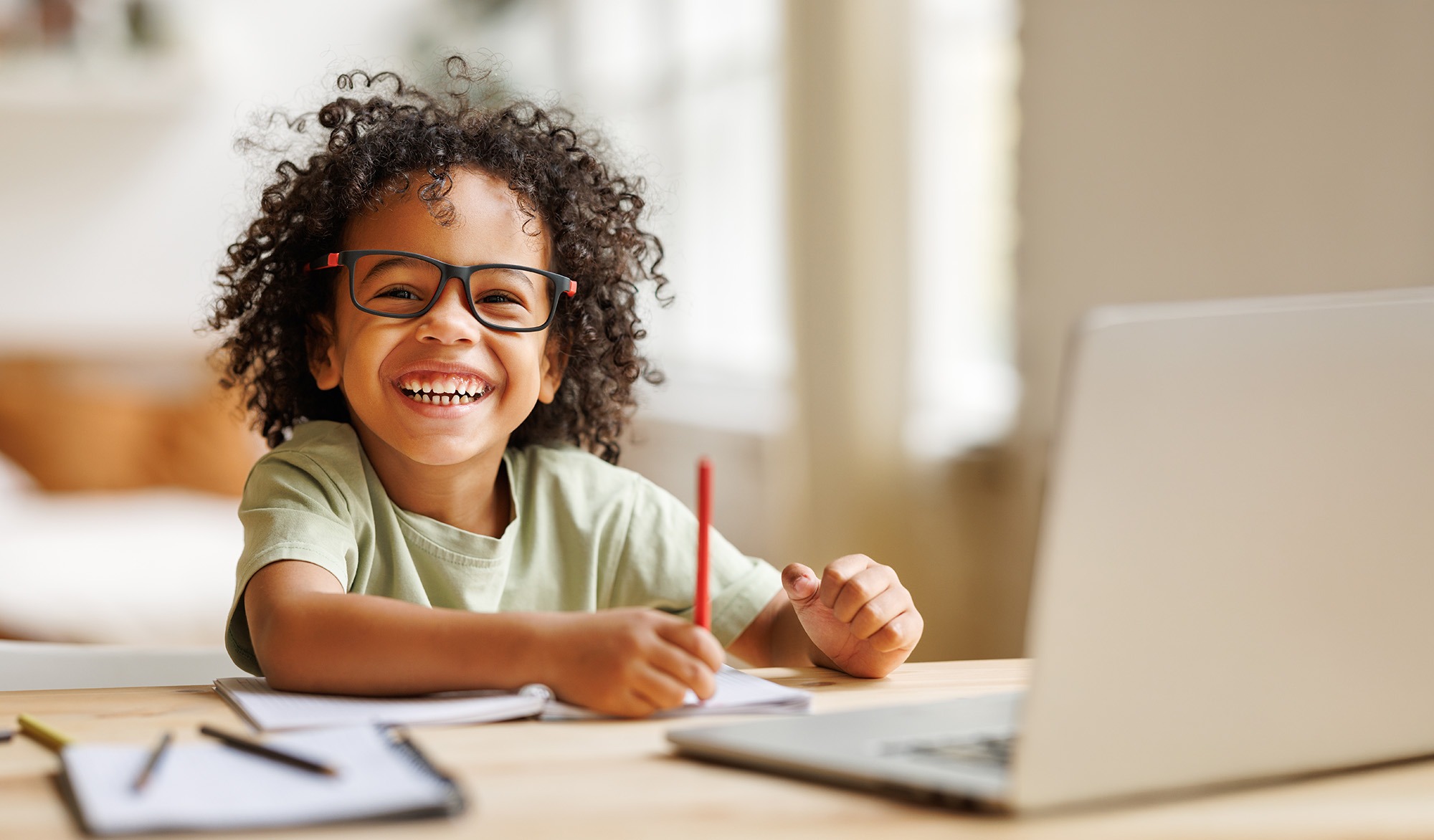 A cheerful child with curly hair and glasses smiles while writing in a notebook at a desk, laptop nearby, highlighting a fun learning environment.