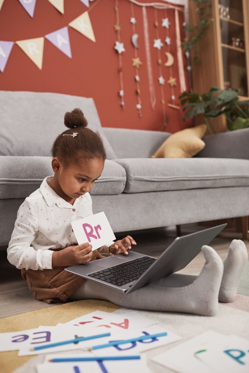 Child using laptop for online learning with alphabet cards at home.